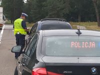 Policjant sprawdza dokumenty kierowcy, z tyłu nieoznakowany samochód policyjny z wyświetlonym napisem POLICJA.