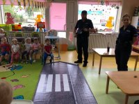 zdjęcie przedstawia kolorową salę przedszkolną. Na podłodze ułożona jest mata z wzorem oznakowanego przejścia dla pieszych. Obok niej stoi umundurowany policjant i policjantka.