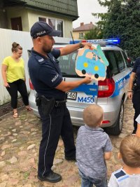 Na zdjęciu widać policjanta na tle radiowozu, oraz kilkoro dzieci. Policjant w dłoniach trzyma laurkę w kształcie radiowozu.