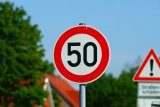 zdjęcie przedstawia znak ograniczenia prędkości do 50 km/h ustawiony przy drodze. Za znakiem widać drogę i budynki.