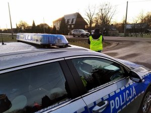 Policjant stoi przy drodze, obok niego stoi radiowóz