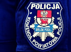 Naszywka na mundurze z napisem POLICJA, KOMENDA POWIATOWA POLICJI BIELSK PODL., odznaką policyjną i herbem powiatu bielskiego.