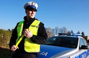 Policjant w mundurze stoi przy radiowozie, w ręku trzyma opaski odblaskowe.