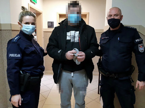 Trzy osoby stoja w budynku, jedna z nich stojąca pośrodku ma załozone na ręce kajdanki, po obu jej stronach stoją policjant i policjantka w mundurach.