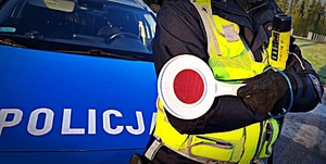Policjant stoi, trzyma tarczkę do zatrzymywania pojazdów i urządzenie do badania stanu trzeźwości, napis POLICJA na radiowozie.