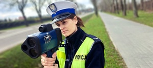 Policjantka w mundurze stoi przy drodze, trzyma w ręku miernik prędkości.