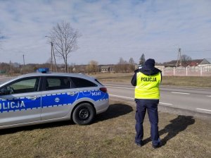 Policjant w mundurze stoi przy drodze, obok niego stoi radiowóz.