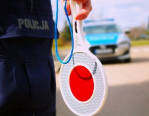 Tarczka do zatrzymywania pojazdów i część umundurowania policjanta, w tle radiowóz policyjny.