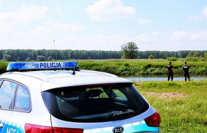 Radiowóz policyjny stoi, dalej widać dwóch policjantów.