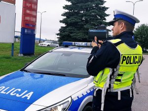 Policjant stoi na świeżym powietrzu, trzyma ręczny miernik prędkości, obok stoi radiowóz.