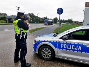 Policjant stoi przy drodze, obok niego stoi radiowóz.