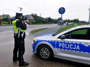 Policjant w mundurze stoi przy drodze, trzyma miernik prędkości, obok stoi radiowóz.