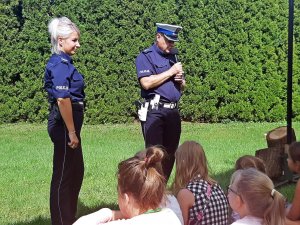 Policjant stoi i trzyma mikrofon, policjantka stoi, dzieci siedza.