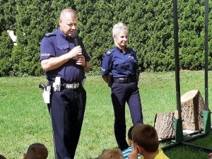 Policjant  i policjantka w mundurach stoją na trawie, przed nimi siedzą dzieci.