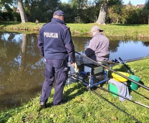 Dwie osoby na świeżym powietrzu nad stawem, jedna jest w mundurze policjanta, druga siedzi i trzyma wędkę.