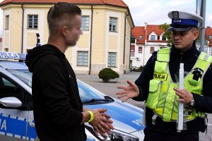 Policjant stoi na świeżym powietrzu, obok stoi mężczyzna, za nimi stoi radiowóz.