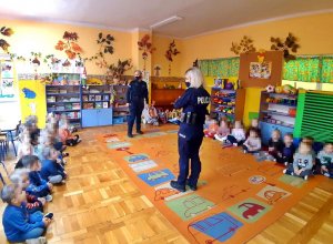 Dwie policjantki w mundurach stoją w sali, na podłodze siedza dzieci.