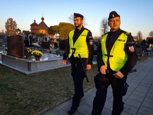 Dwaj policjanci w mundurach idą aleją na cmentarzu.
