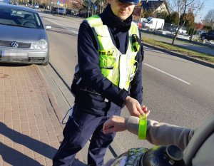 Policjant w mundurze stoi i zakłada element odblaskowy na rękę innej osoby.