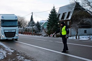 Policjant w mundurze stoi przy drodze, drogą jedzie samochód ciężarowy.