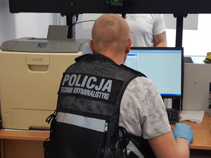 Mężczyzna siedzi przy biurku, ma załozona kamizelkę z napisem POLICJA TECHNIK KRYMINALISTYKI