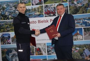 Dwaj mężczyźni stoją w budynku i podają sobie ręce, za nimi widać zdjęcia Bielska Podlaskiego.