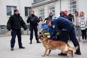 Policjant, osoby i pies stoją na placu komendy.